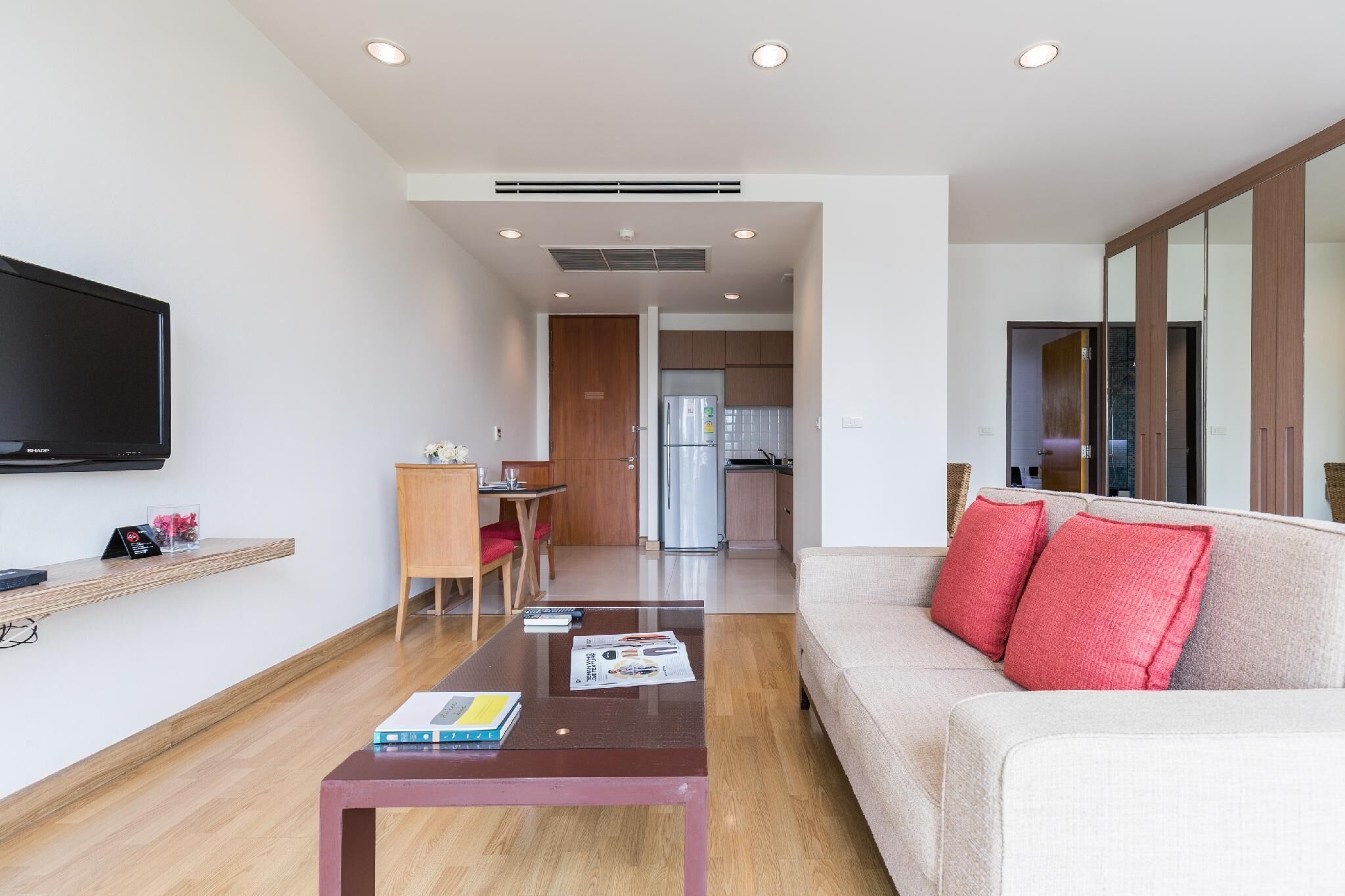 A modern living room with a sofa, coffee table, and a flat-screen TV. There is a kitchen area in the background with a refrigerator and cabinets.