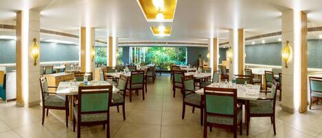 A spacious dining area with multiple tables and chairs, large windows with a view of greenery, and modern lighting fixtures.
