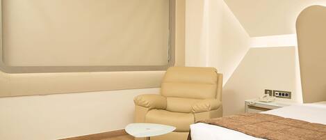 Une chambre d’hôtel avec une grande fenêtre, un fauteuil beige, une petite table blanche, un lit recouvert d’un plaid marron et un plancher en bois.
