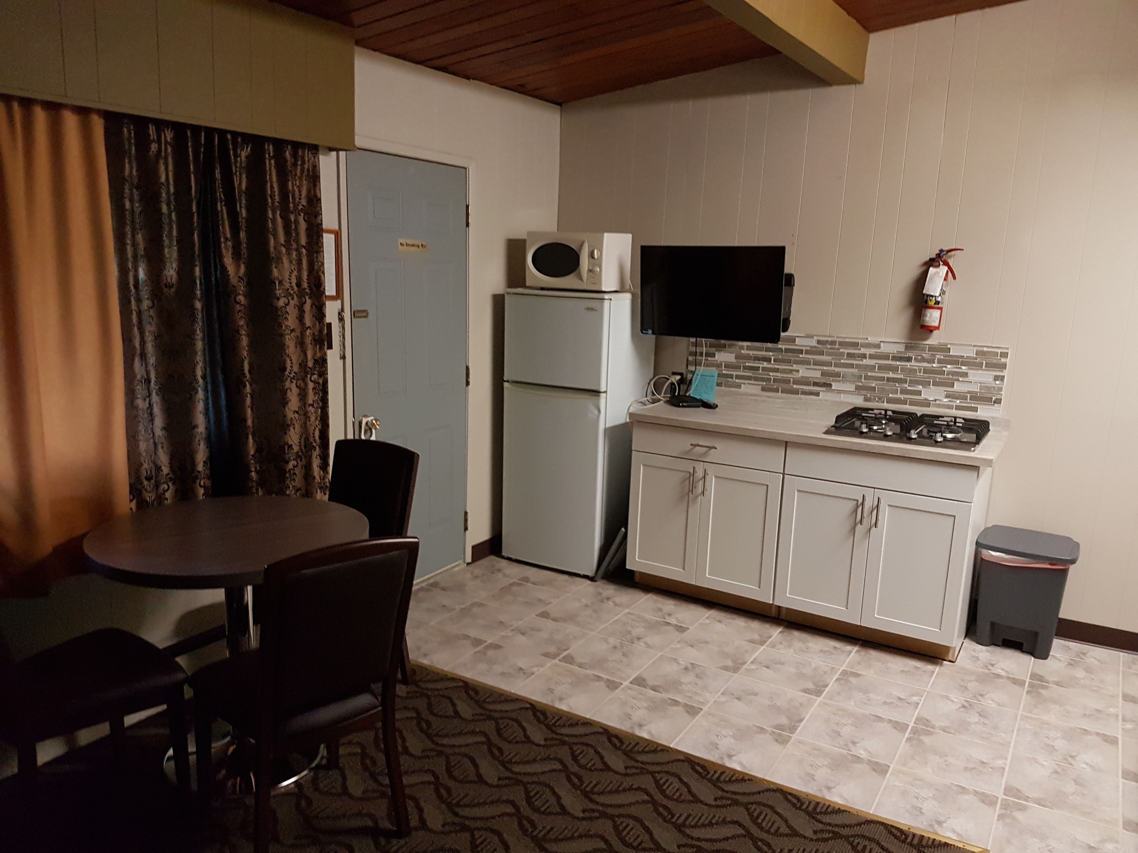 A compact kitchen with a refrigerator, microwave, stove, and a flat-screen TV. There is a dining table with chairs and a patterned rug.