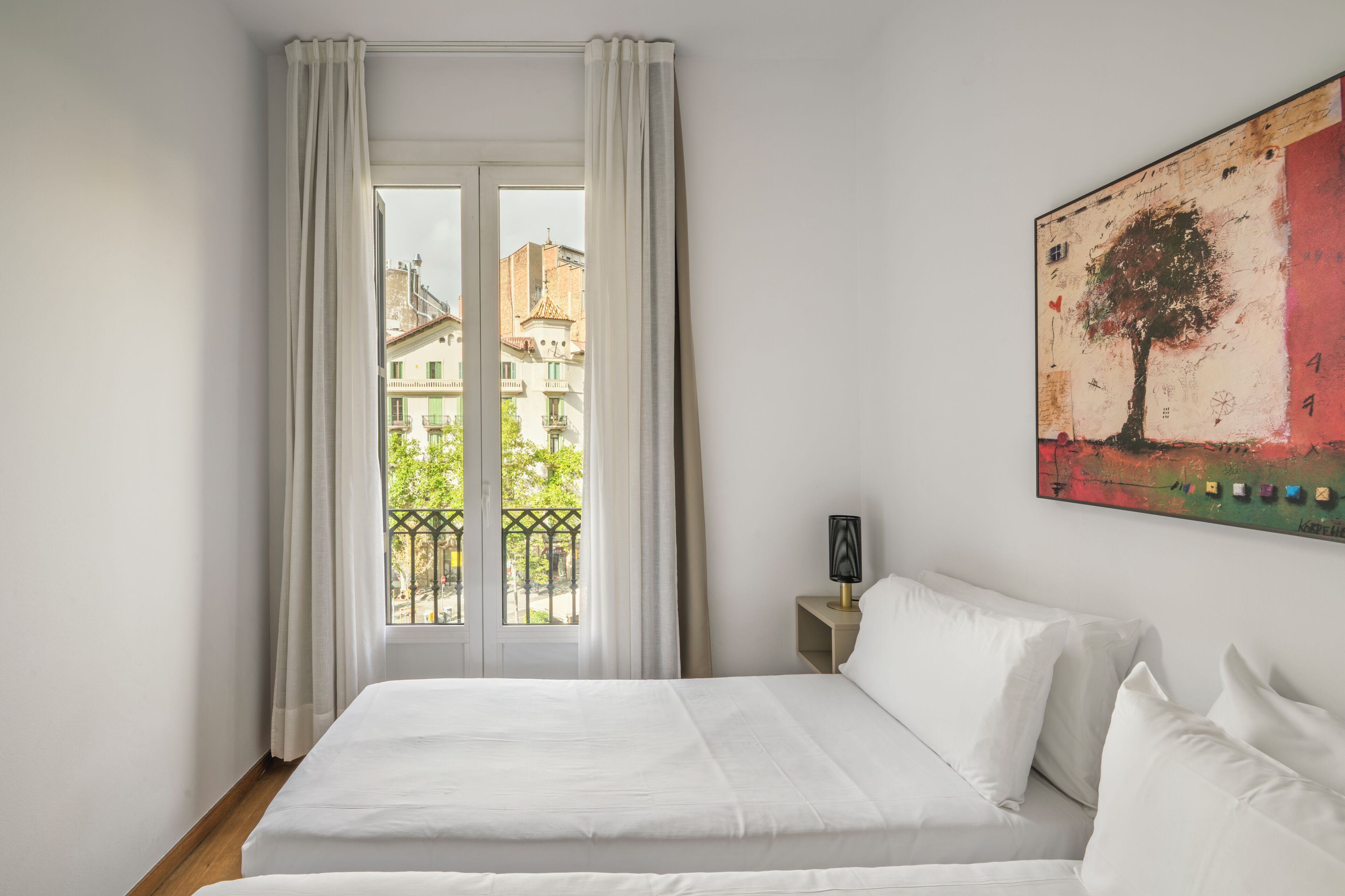 Un dormitorio con una cama grande, una ventana con cortinas, una mesita de noche con lámpara y un cuadro en la pared.