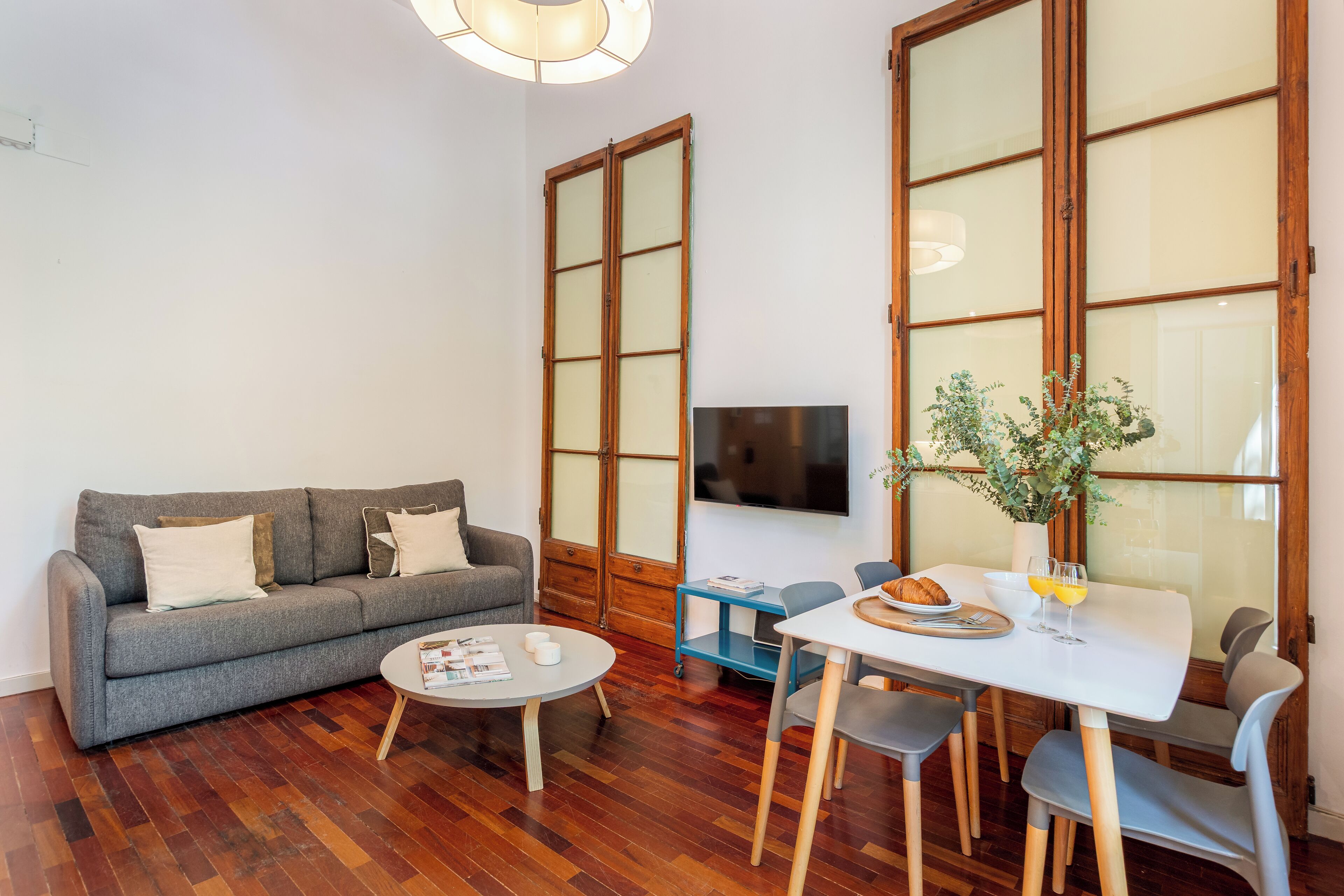 A living room with a grey sofa, a round coffee table, a dining table with chairs, a TV on the wall, and a large window with wooden frames.
