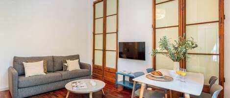 A living room with a grey sofa, a round coffee table, a dining table with chairs, a TV on the wall, and a large window with wooden frames.