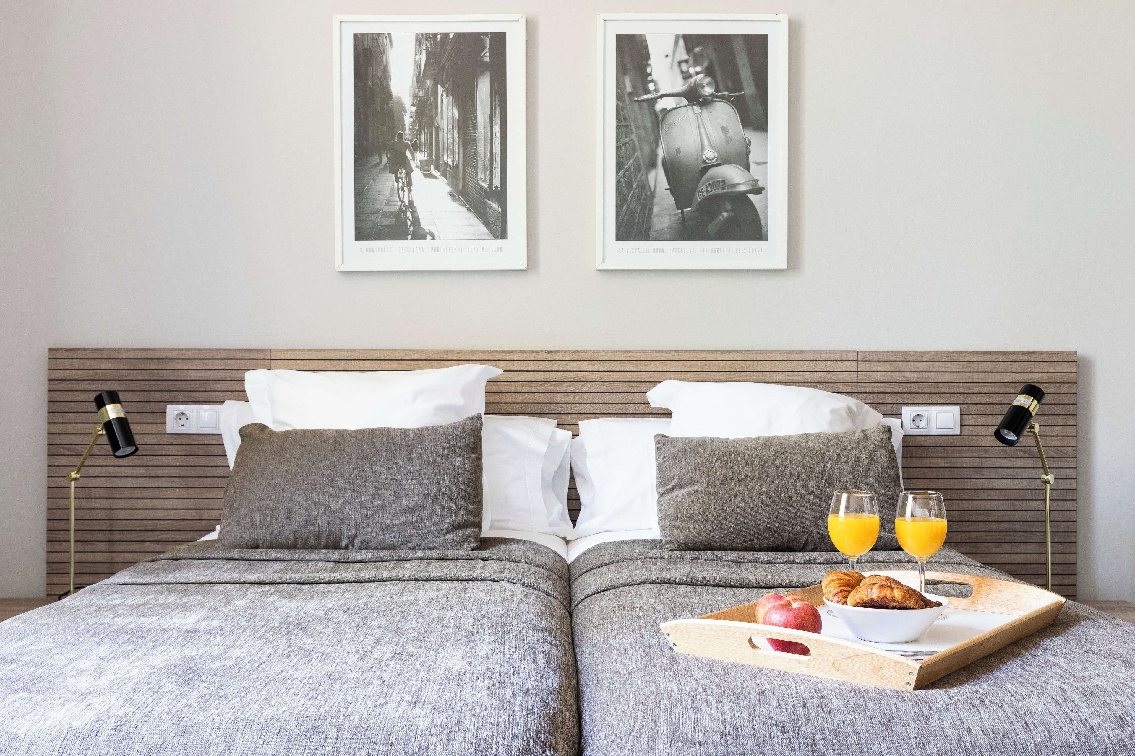 Un lit double avec deux oreillers, une table de chevet avec une lampe, et un plateau avec des croissants, des fruits et du jus d’orange.
