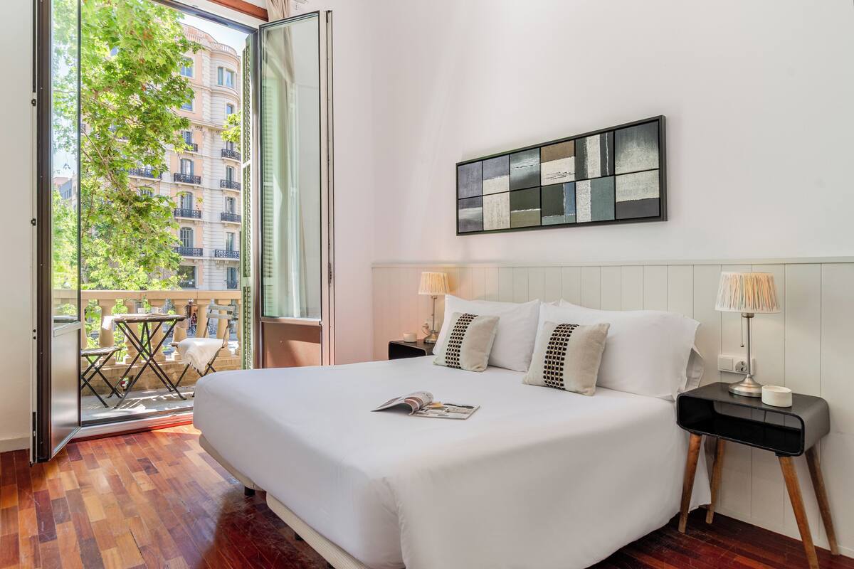 Habitación de hotel con una cama grande, una mesita de noche, una lámpara, una revista sobre la cama y vistas a un edificio a través de la puerta abierta.
