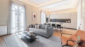 A modern living room with a grey sofa, a coffee table, and a brown armchair. There is a dining area with a table and chairs, and a kitchen in the background. - Aspasios Rambla Catalunya Suites  (Barcelona)