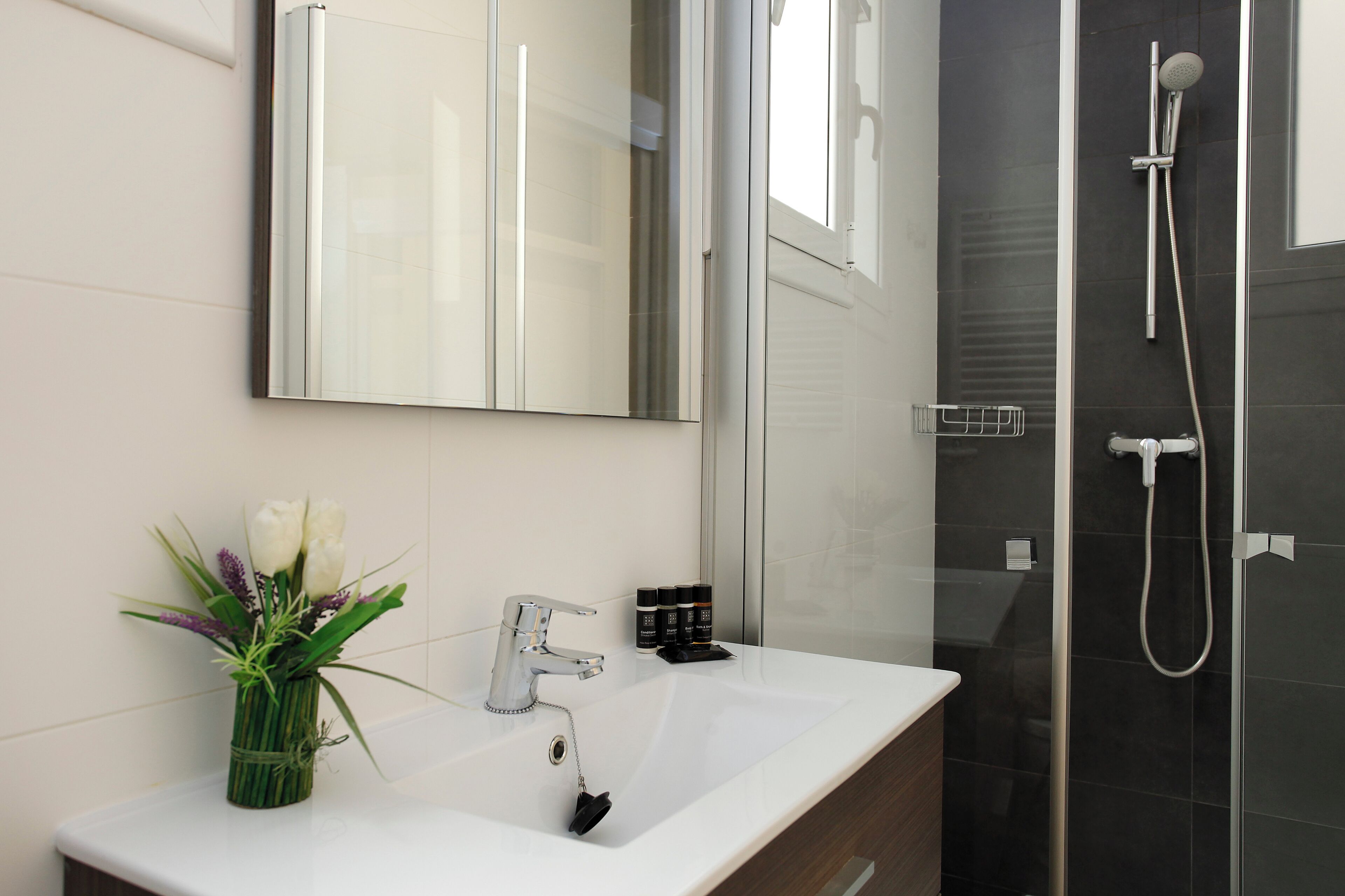 A bathroom with a white sink, a mirror, a shower, and a vase with flowers.