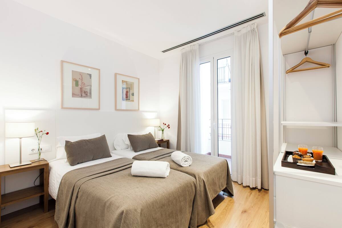 Habitación de hotel con dos camas, una mesita de noche con lámpara, una pequeña mesa con bandeja de desayuno y un perchero.