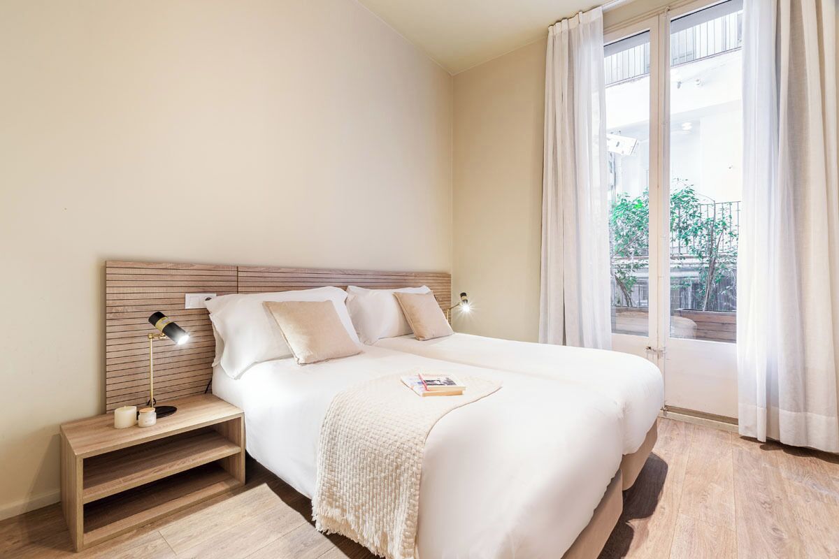 Una cama bien tendida con una colcha blanca y almohadas, una mesita de noche con una lámpara y una ventana con cortinas.