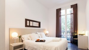 A neatly arranged hotel room with a large bed, bedside tables with lamps, a small table with a tray of items, and a view through a window with curtains. - Aspasios Rambla Catalunya Suites  (Barcelona)