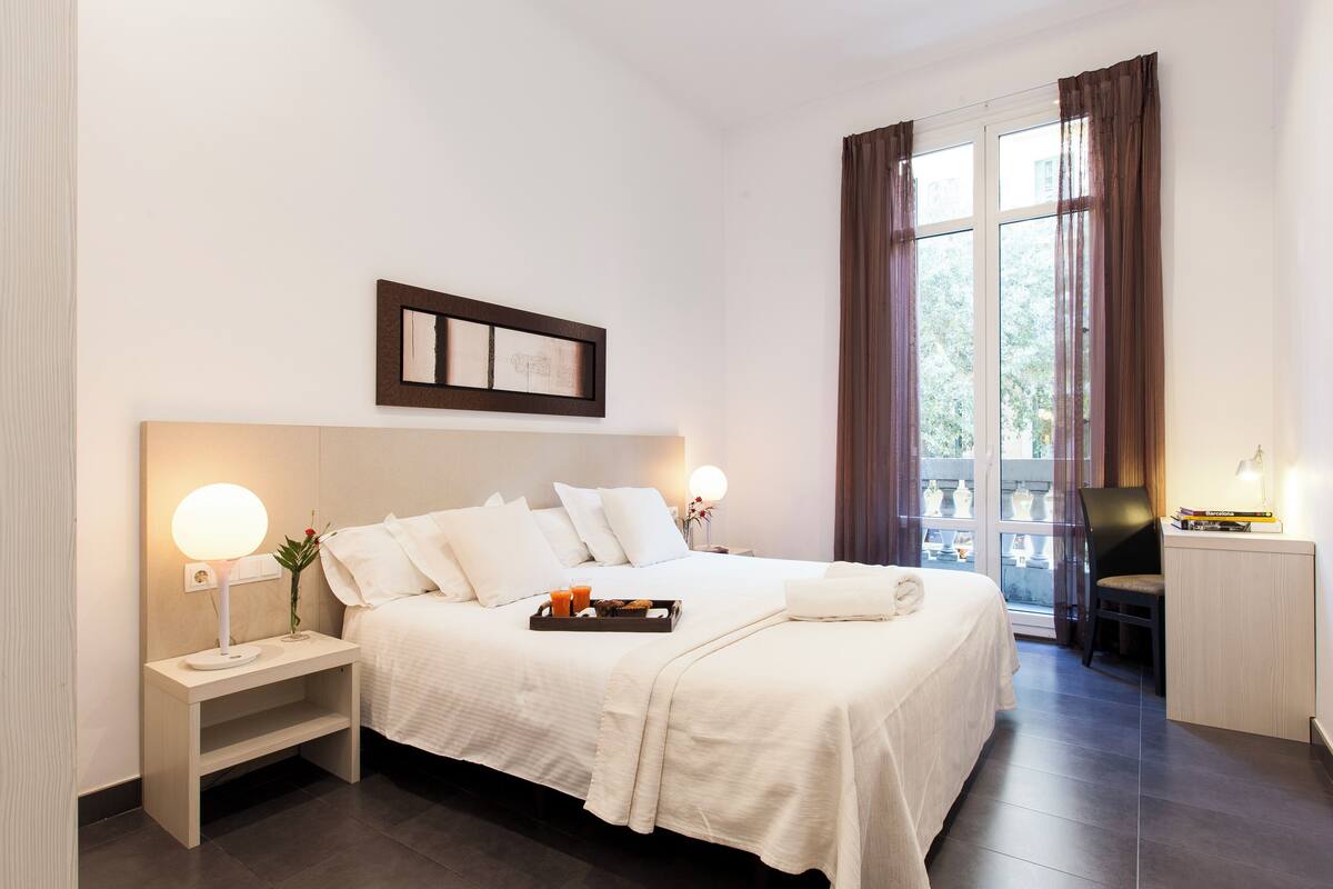 Habitación de hotel ordenada con una cama grande, mesitas de noche con lámparas, una pequeña mesa con una bandeja de objetos y vistas a través de una ventana con cortinas.