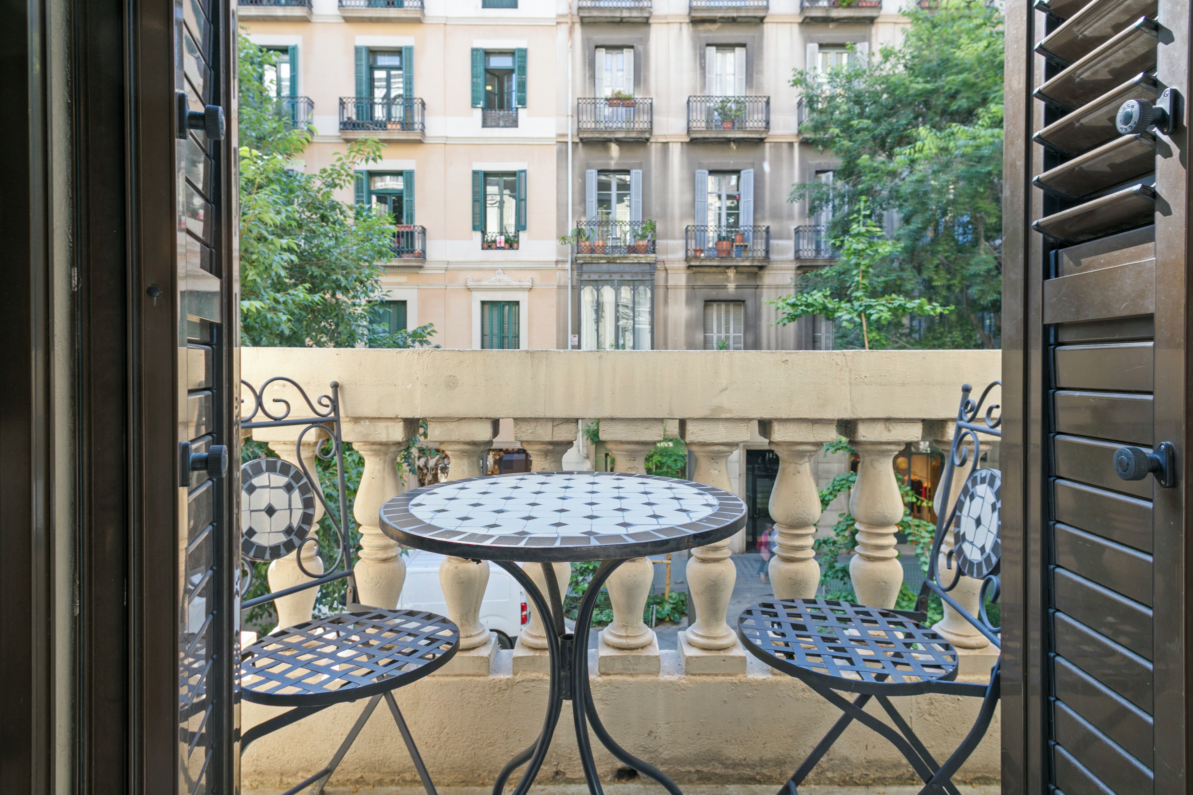 An open balcony with a round table and two chairs, overlooking a building with balconies and greenery.