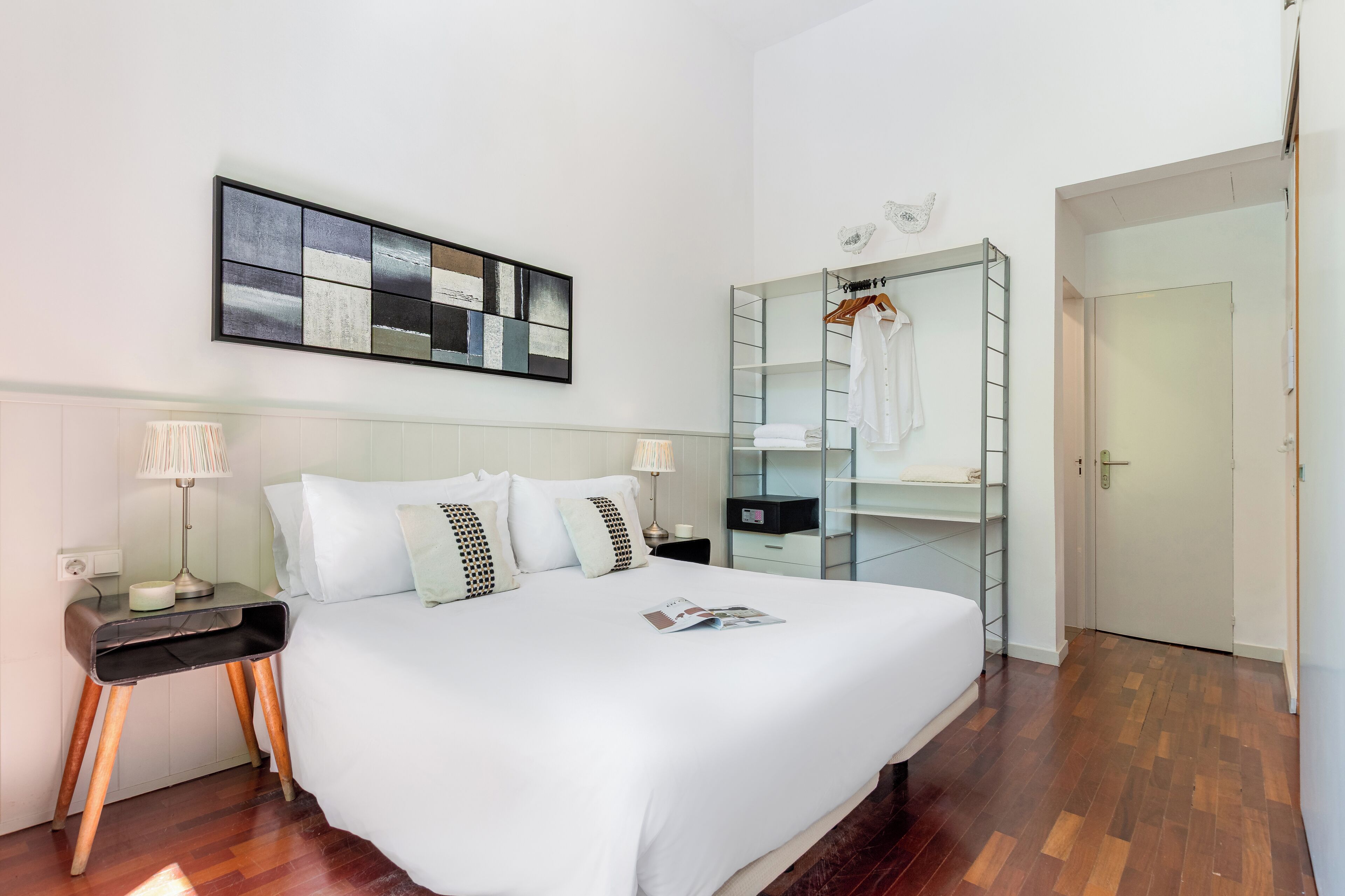A modern hotel room with a large bed, a bedside table with a lamp, a wall-mounted artwork, a wardrobe with a mirror, and a door leading to another room.