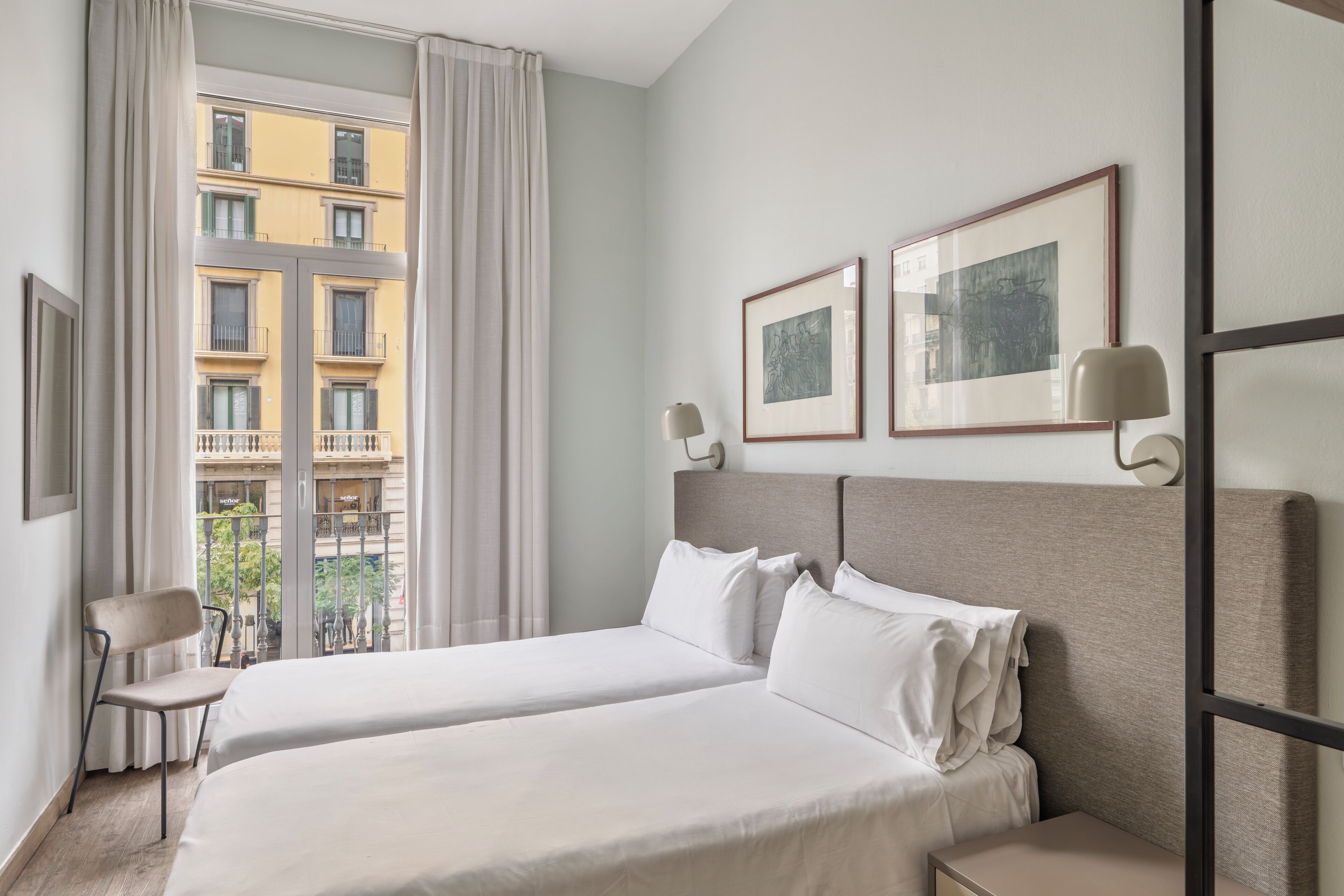 Ein Hotelzimmer mit zwei Betten, einem Sessel, einem Nachttisch und einem Fenster mit Blick auf ein Gebäude.
