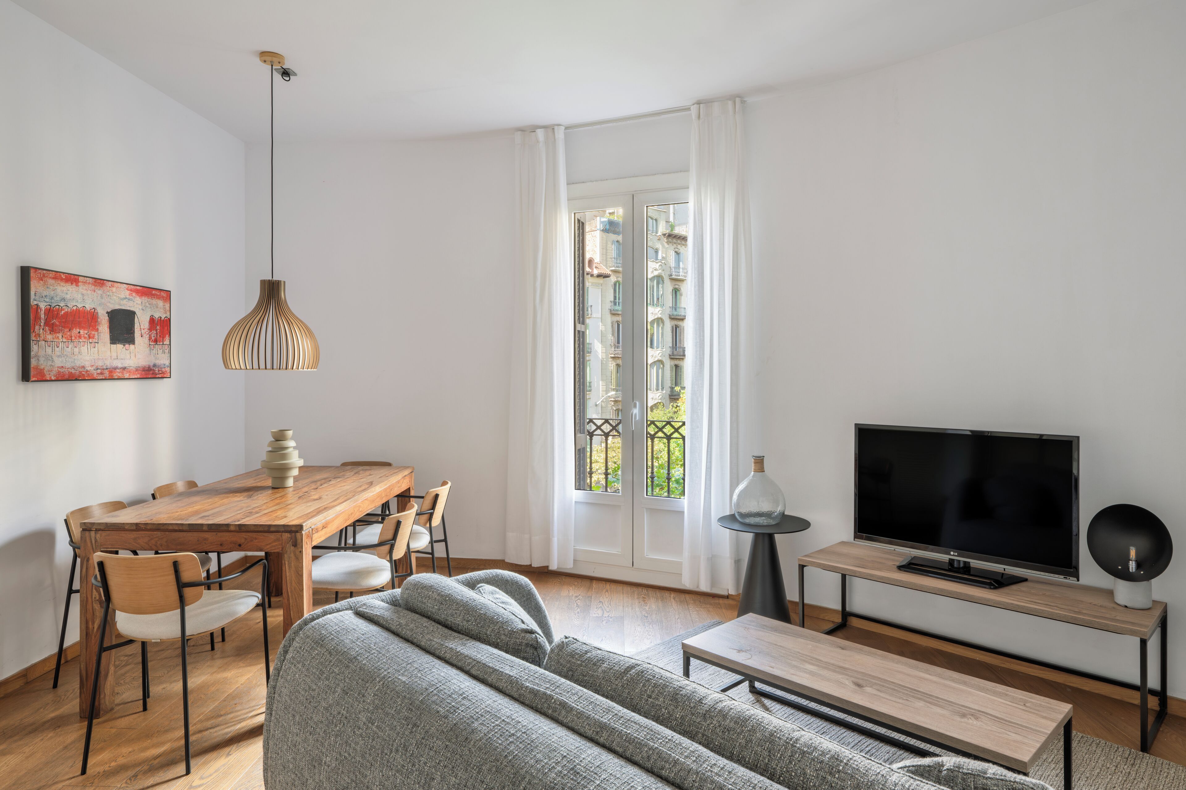 A modern living room with a wooden dining table, grey sofa, and a television on a wooden stand.