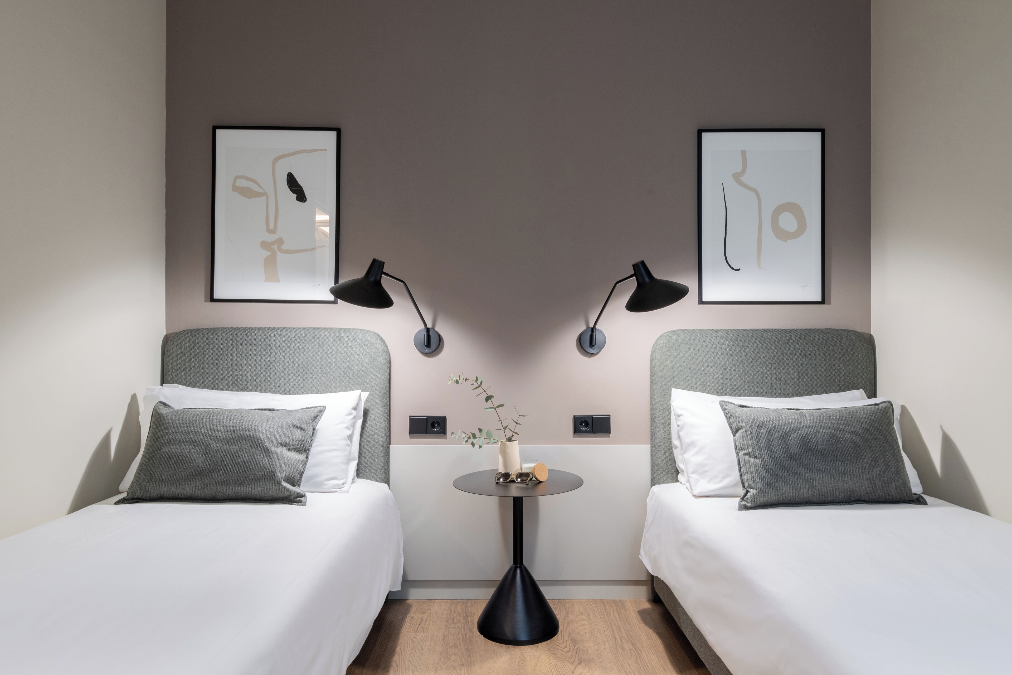 Habitación de hotel con dos camas, una mesita redonda con un florero y dos lámparas de pared.