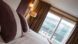 A hotel room with a large bed, a nightstand with a lamp, and a view of a mountainous landscape through the window. - Cairndow Stagecoach Inn (Cairndow)