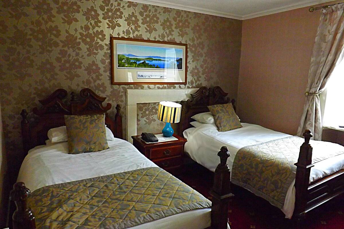 A hotel room with two beds, a nightstand with a lamp, a framed picture on the wall, and a window with curtains.