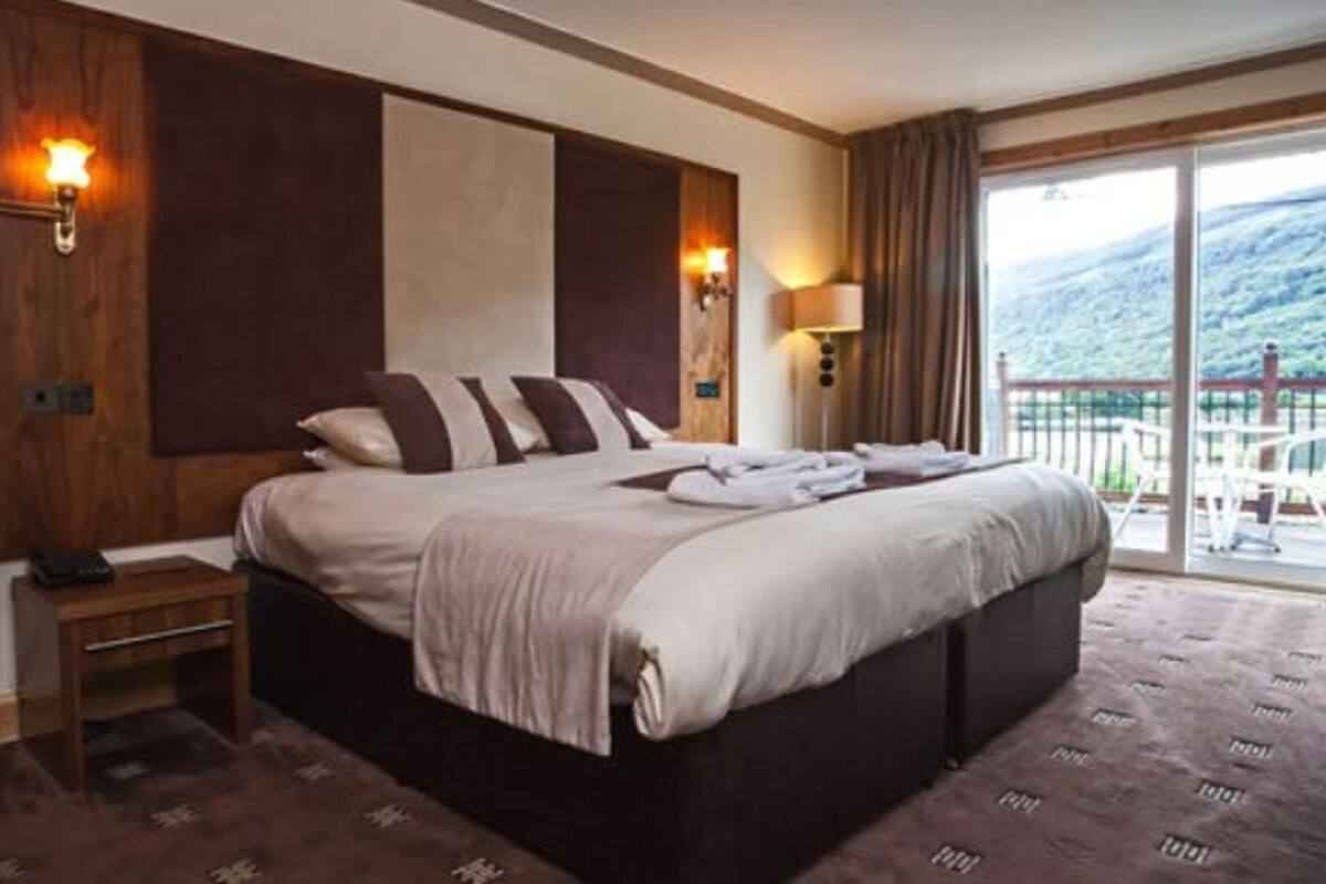 A hotel room with a large bed, a wooden headboard, a nightstand, a lamp, and a view of a balcony with outdoor furniture.