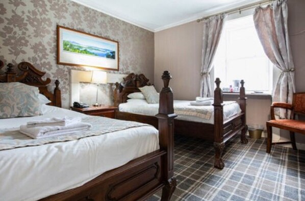 A room with two wooden beds, a chair, a nightstand, a lamp, and a window with curtains.