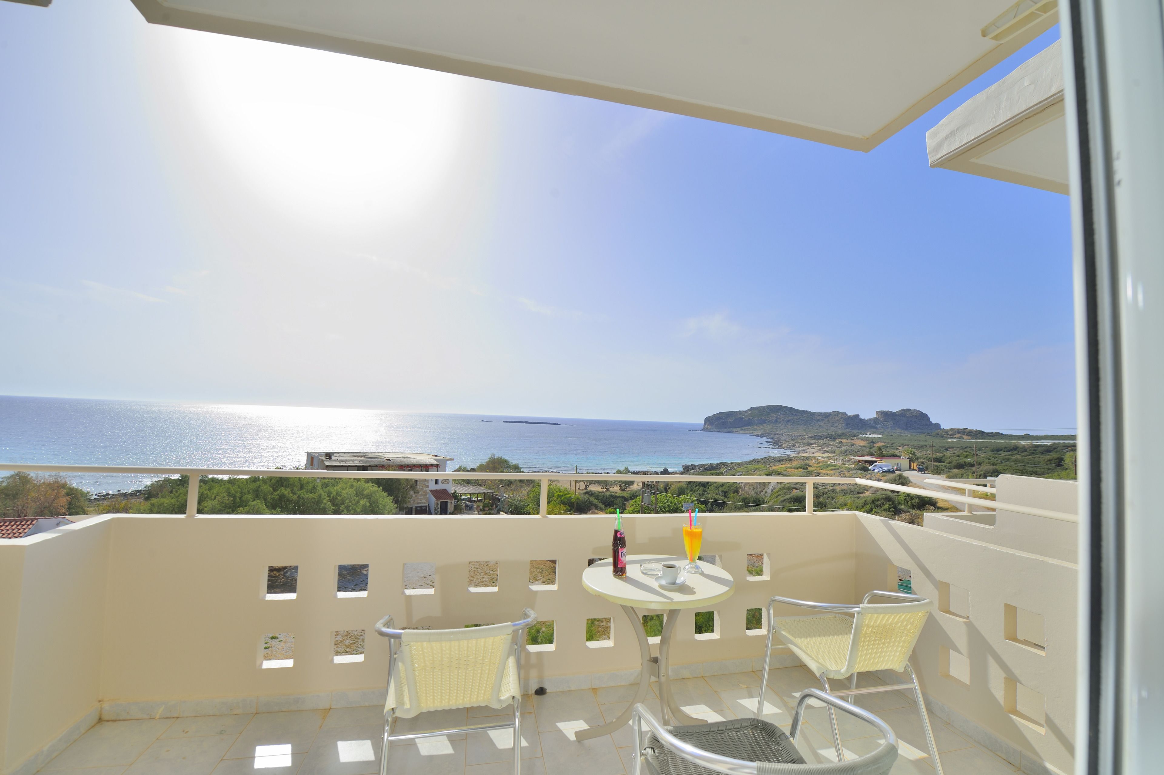 Un balcón con vista al mar, sillas blancas y una mesa con bebidas.