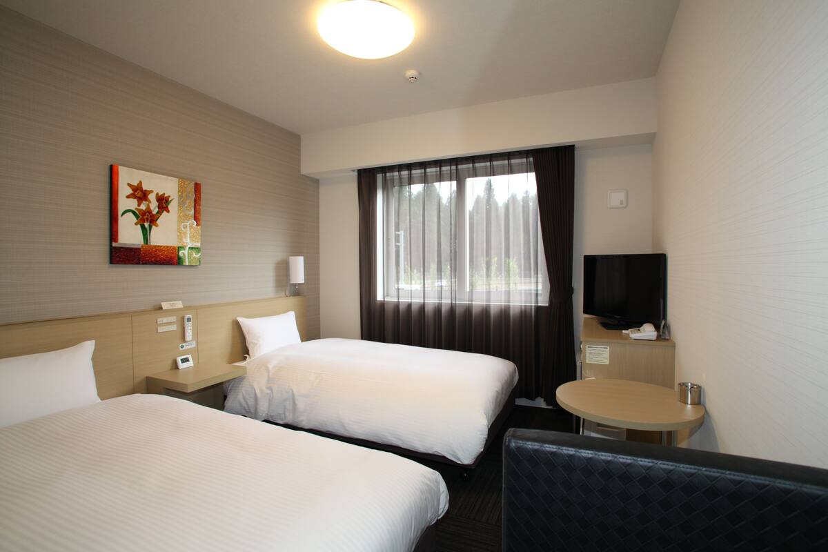 A hotel room with two beds, a television, a small table, and a window with curtains.
