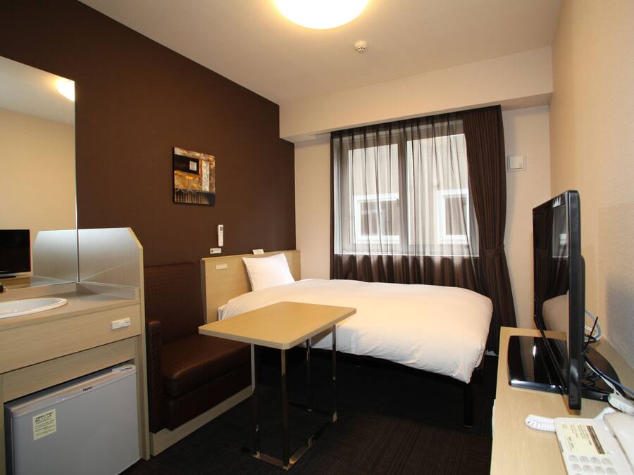 A hotel room with a bed, a desk, a chair, a small table, a desk lamp, a television, and a refrigerator.