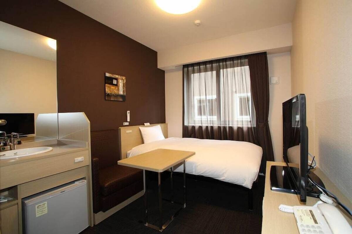 A hotel room with a bed, a desk, a chair, a TV, a phone, and a small refrigerator.