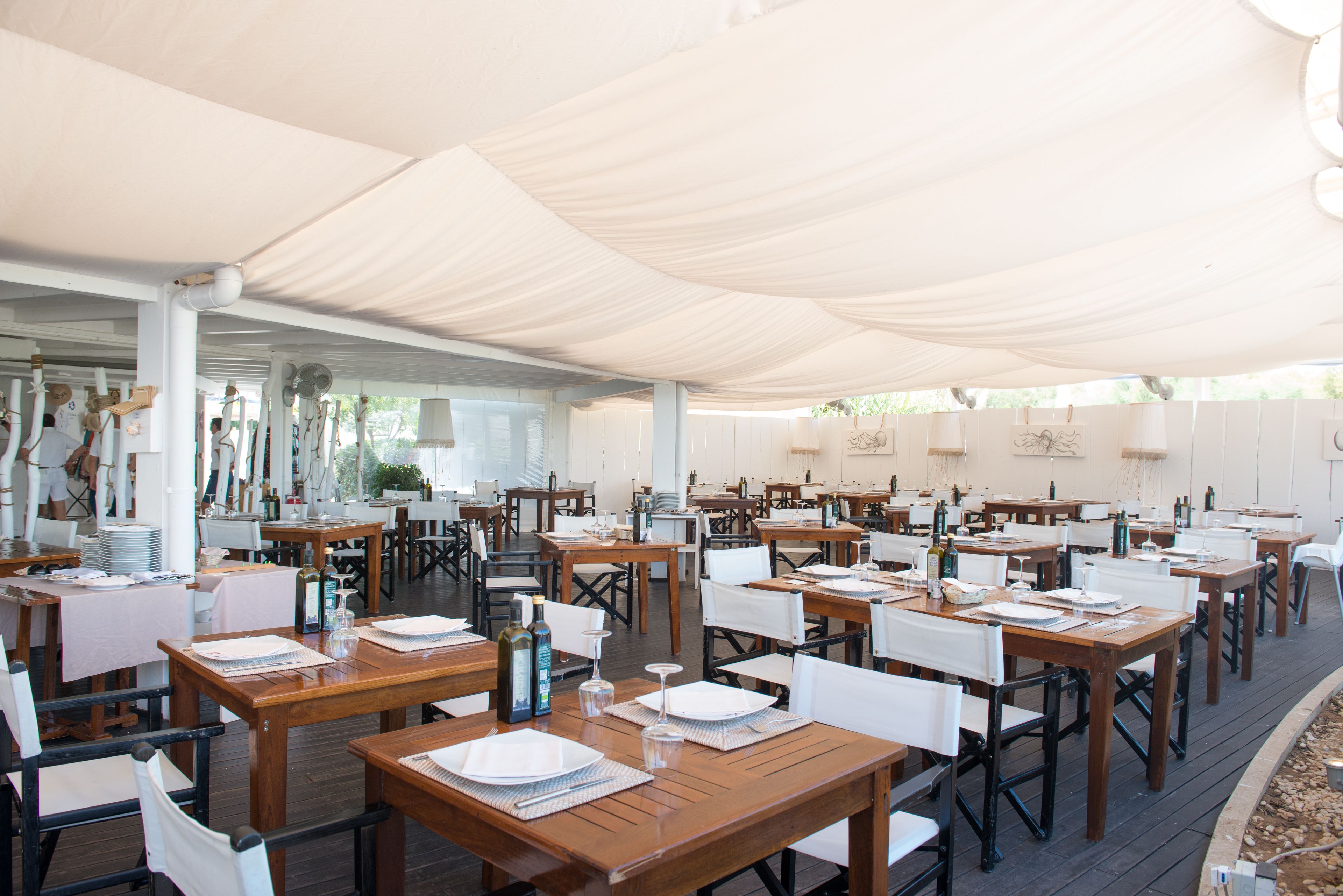 An outdoor dining area with wooden tables and white chairs, covered by a white canopy.