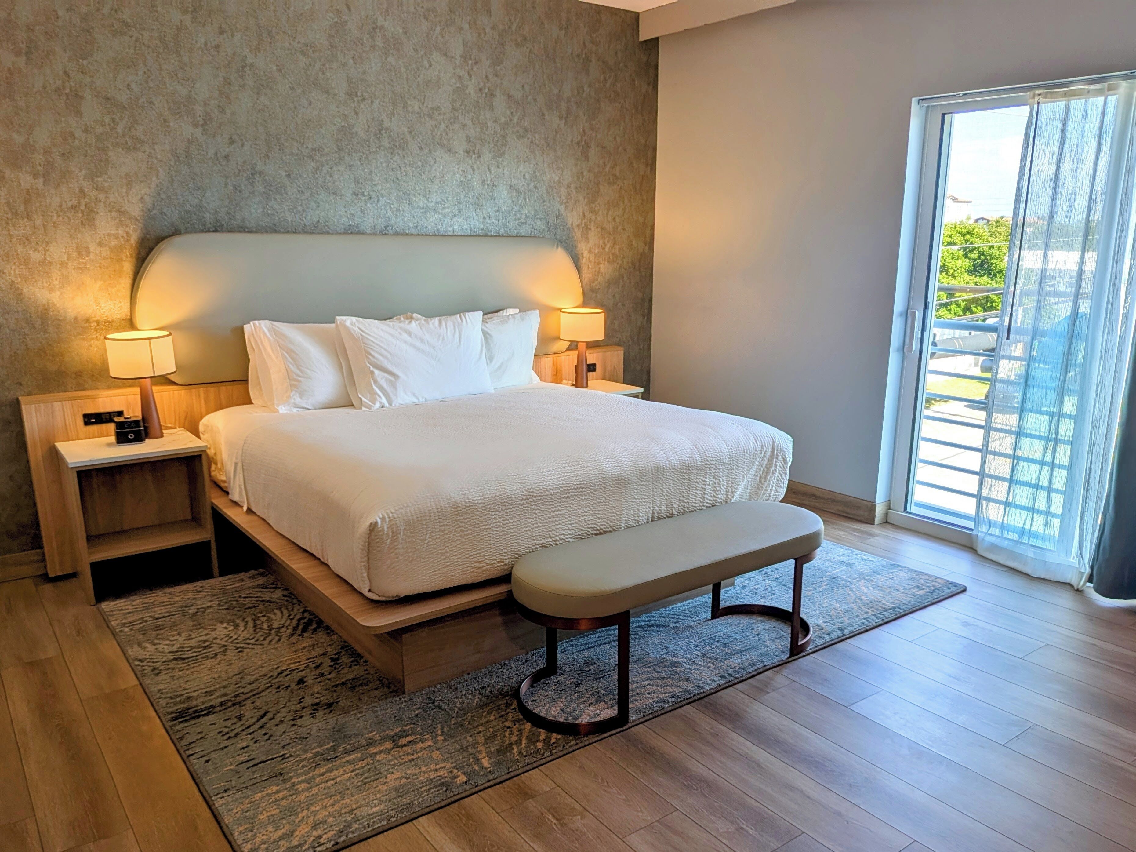 A modern bedroom with a large bed, bedside lamps, a bench, and a view through a sliding glass door.