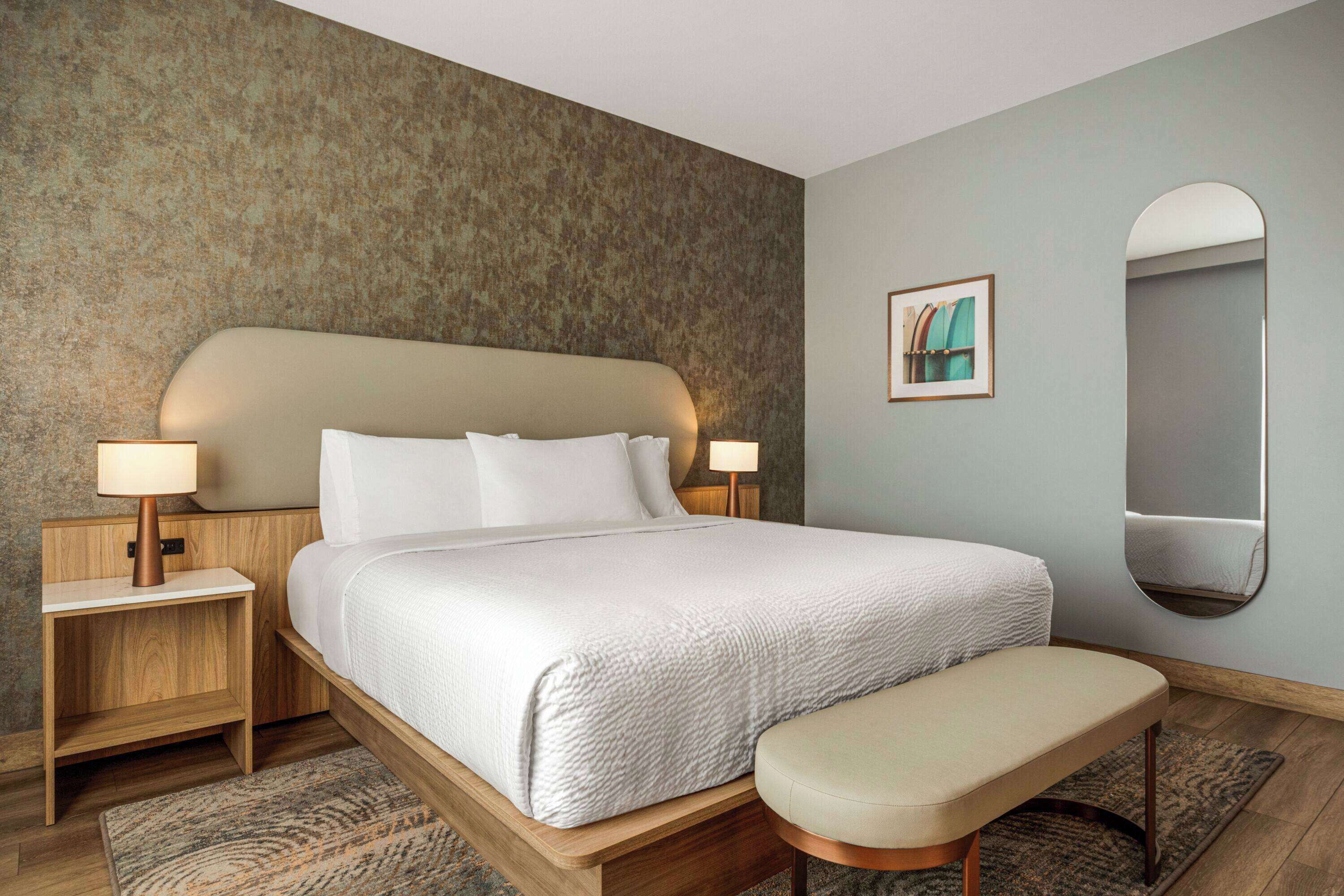 A neatly arranged bedroom with a large bed, bedside lamps, a bench, a mirror, and a framed picture on the wall.