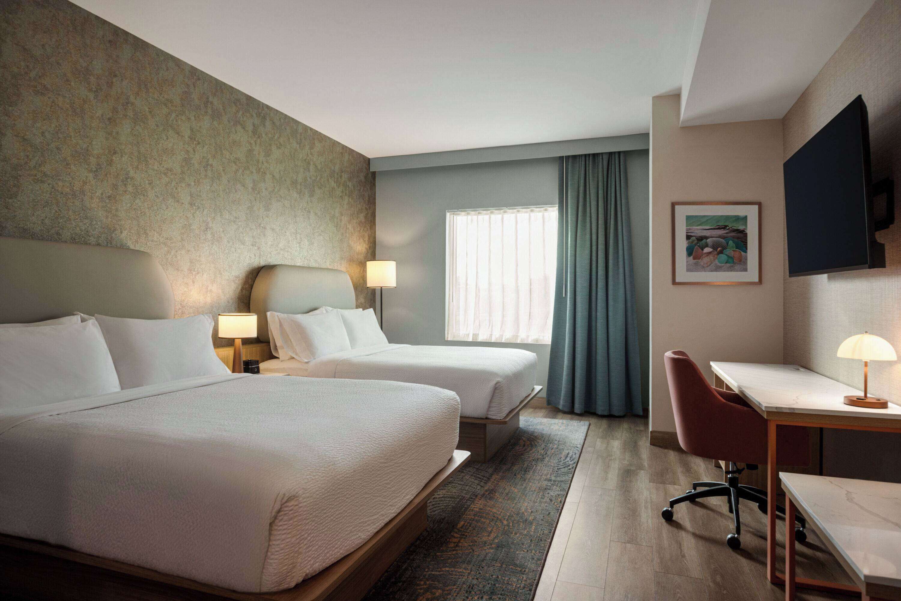 Ein Hotelzimmer mit zwei Betten, einem Schreibtisch, einem Stuhl, einem Fernseher und einem Fenster mit Vorhängen.