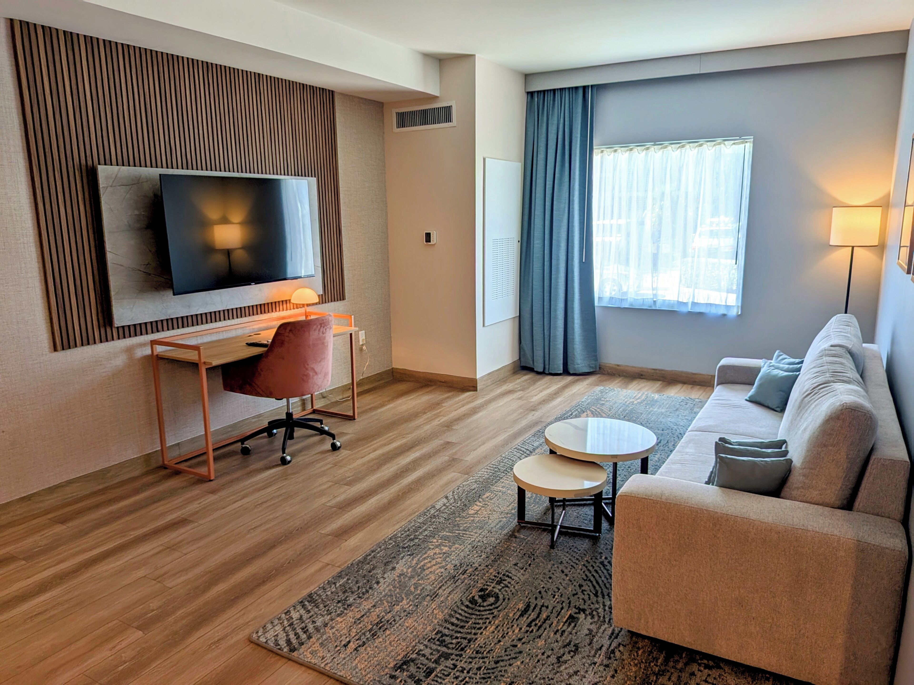 A modern hotel room with a flat-screen TV mounted on the wall, a wooden desk with a chair, a round coffee table, a sofa, and a large window with curtains.
