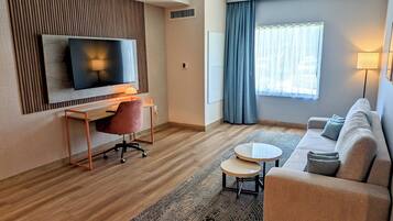 A modern hotel room with a flat-screen TV mounted on the wall, a wooden desk with a chair, a round coffee table, a sofa, and a large window with curtains.