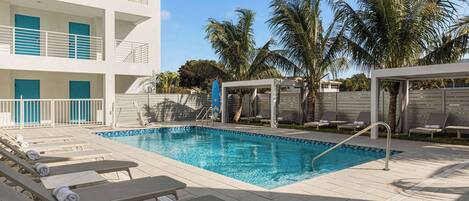 A swimming pool with lounge chairs and a covered seating area, surrounded by palm trees and a multi-story building with balconies.