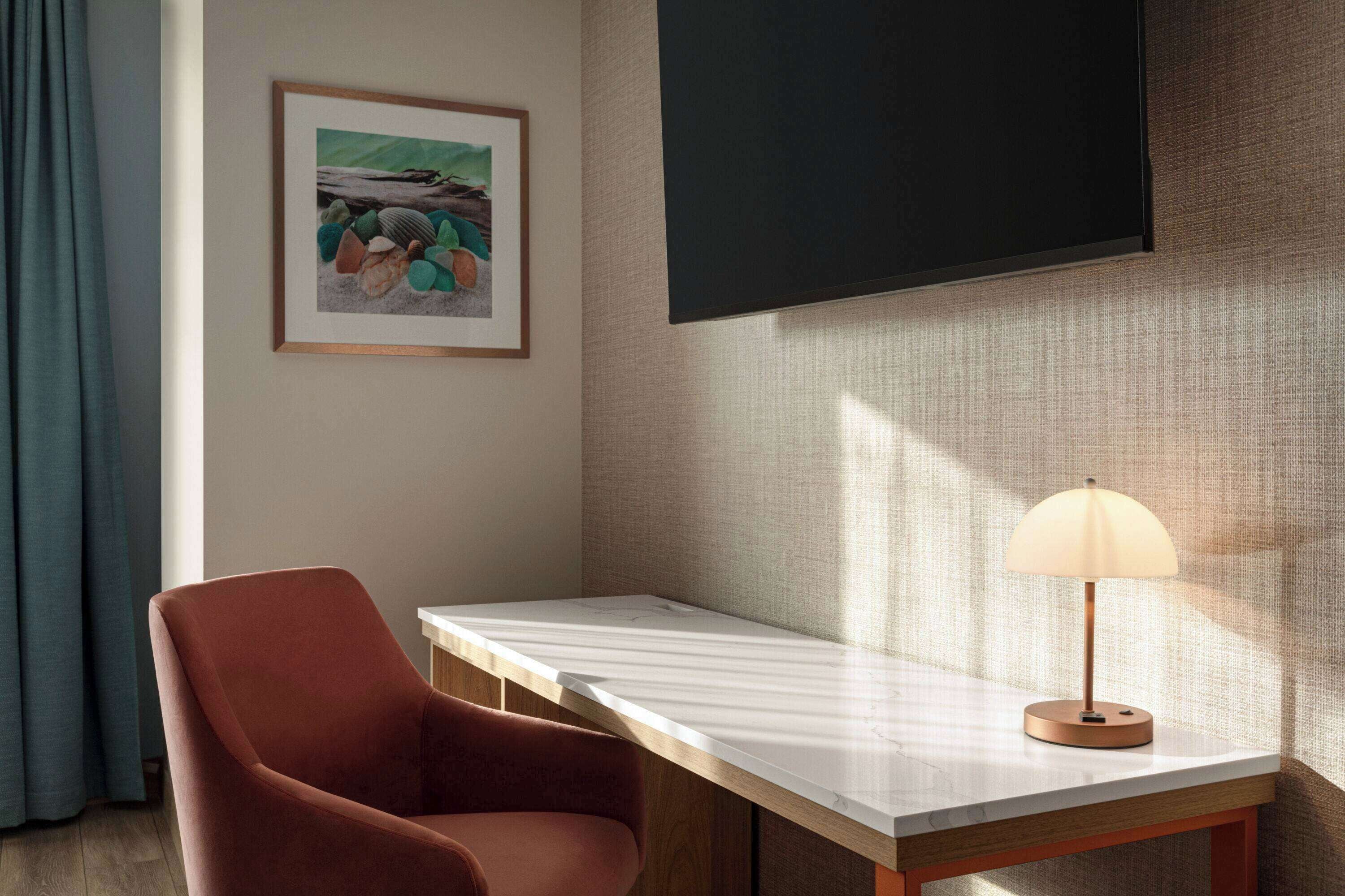 A room with a wooden desk, a cushioned chair, a framed picture on the wall, and a lamp on the desk.