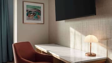 A room with a wooden desk, a cushioned chair, a framed picture on the wall, and a lamp on the desk.