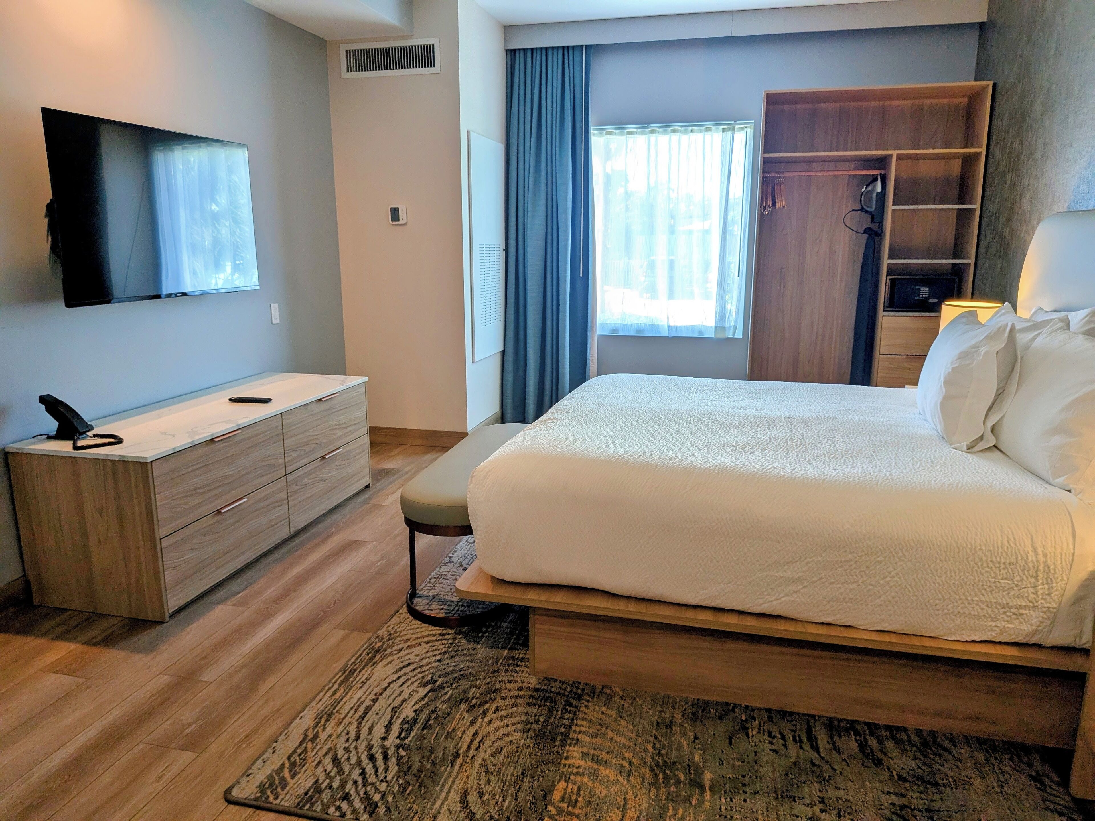 A modern hotel room with a large bed, a wooden dresser, a flat-screen TV mounted on the wall, and a window with curtains.