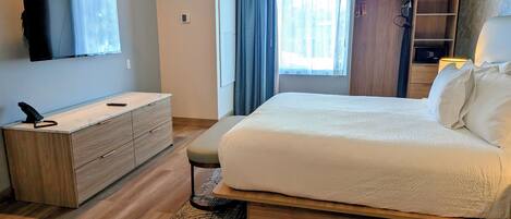 A modern hotel room with a large bed, a wooden dresser, a flat-screen TV mounted on the wall, and a window with curtains.