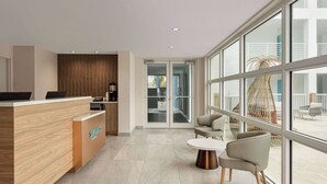 A modern reception area with a wooden reception desk, seating area with chairs and a table, large windows, and a view of the outdoor space. - Sea Glass Beach Place, Trademark Collection by Wyndham (Lauderdale-by-the-Sea)