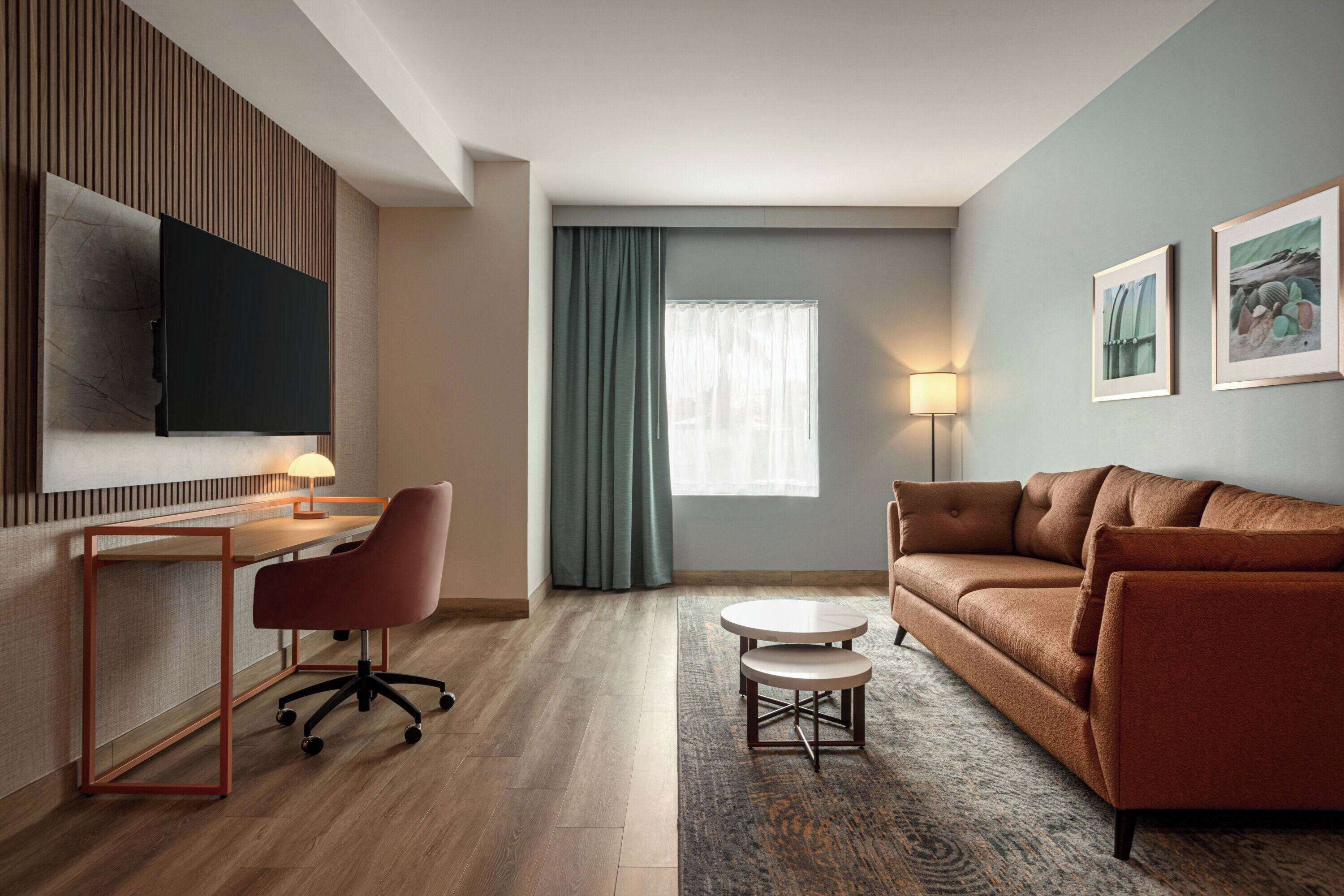 A modern hotel room with a wooden desk, office chair, brown sofa, and a TV mounted on the wall.