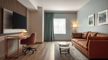 A modern hotel room with a wooden desk, office chair, brown sofa, and a TV mounted on the wall.
