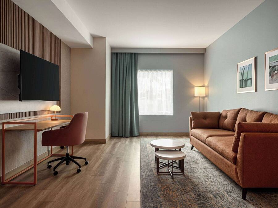 Een moderne hotelkamer met een houten bureau, bureaustoel, bruine bank en een aan de muur bevestigde televisie.