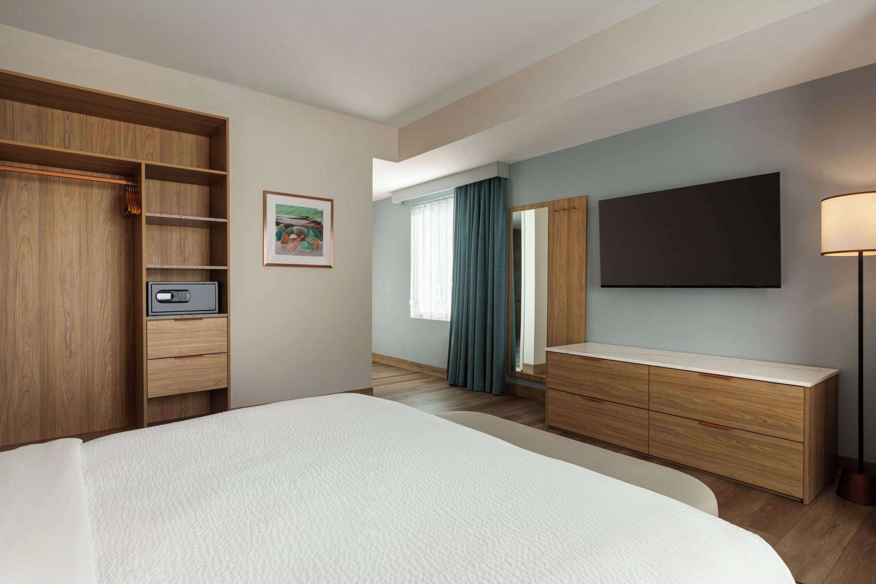 A hotel room with a large bed, wooden wardrobe, a flat-screen TV, a bedside table, and a window with curtains.