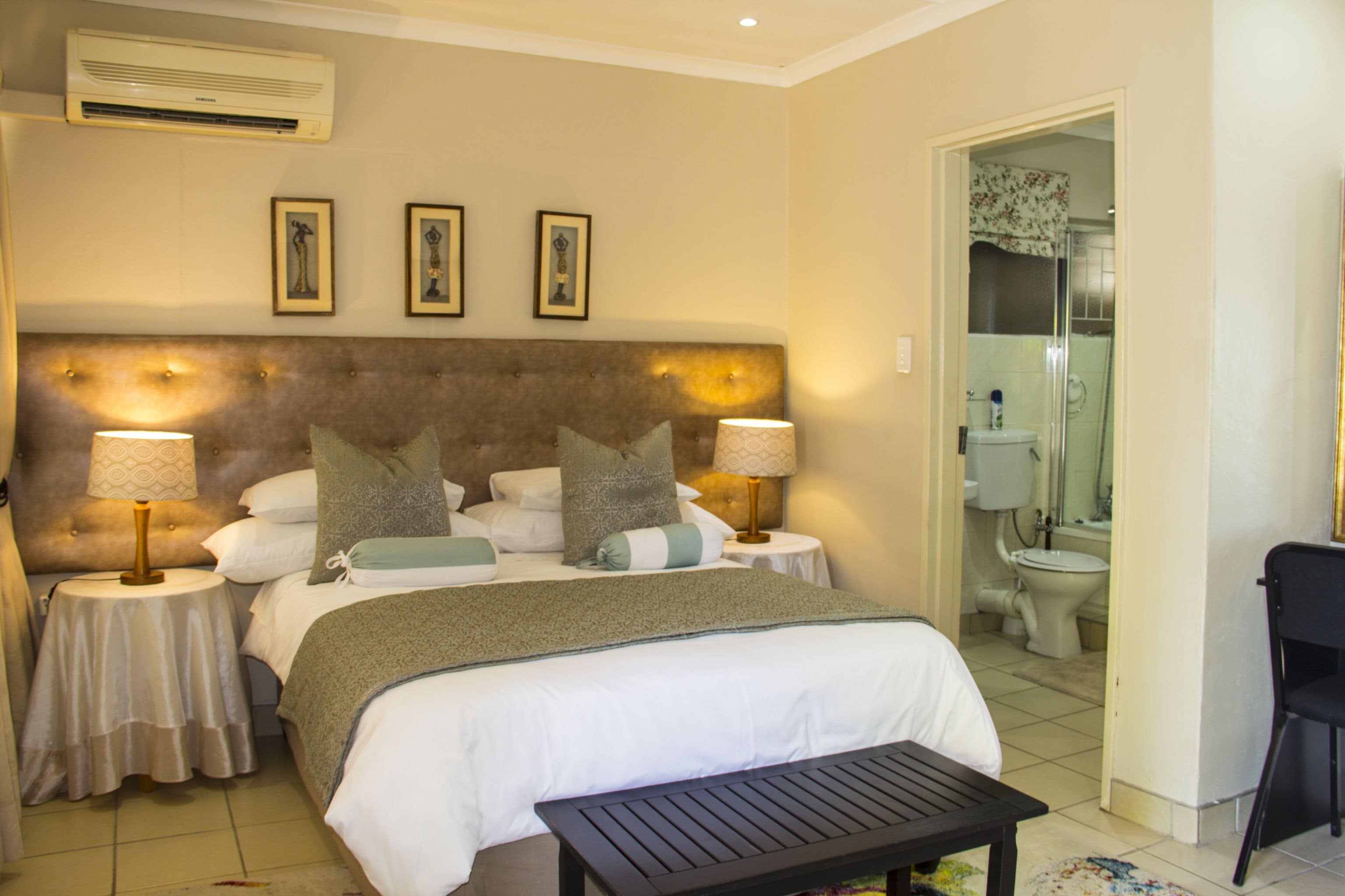 A hotel room with a large bed, two bedside tables with lamps, a small table, and a shower area visible through an open door.