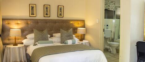 A hotel room with a large bed, two bedside tables with lamps, a small table, and a shower area visible through an open door.