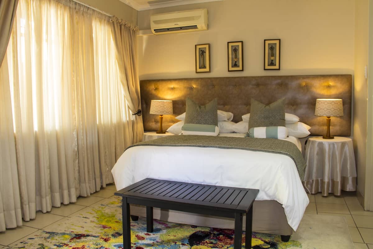 A well-lit hotel room with a large bed, two bedside tables with lamps, a small table, and a colorful rug.