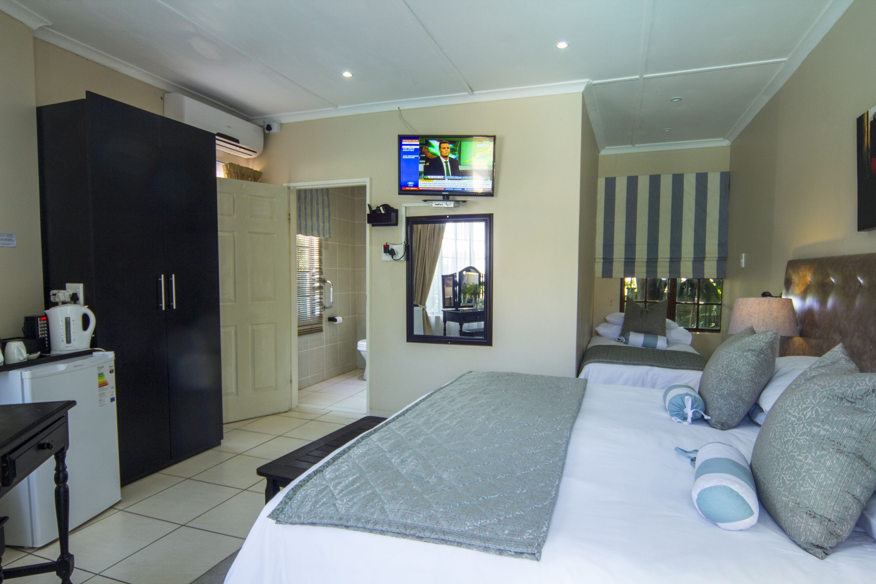 Une chambre d’hôtel avec un lit, un téléviseur fixé au mur, une armoire, un bureau avec une bouilloire et une salle de bain visible à travers une porte ouverte.