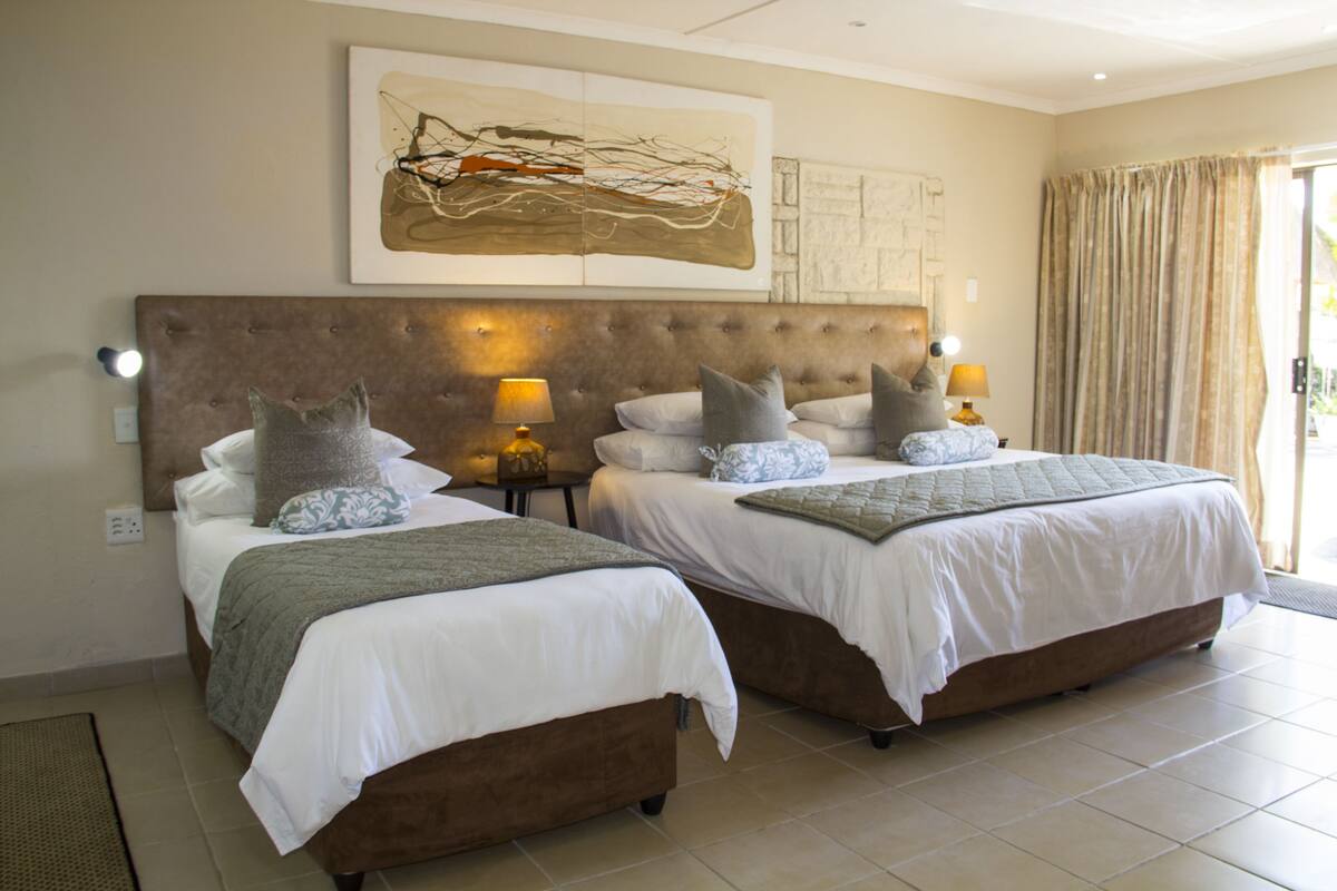 A hotel room with two beds, each with a headboard, white bedding, and multiple pillows. There are bedside lamps and a large abstract painting on the wall.