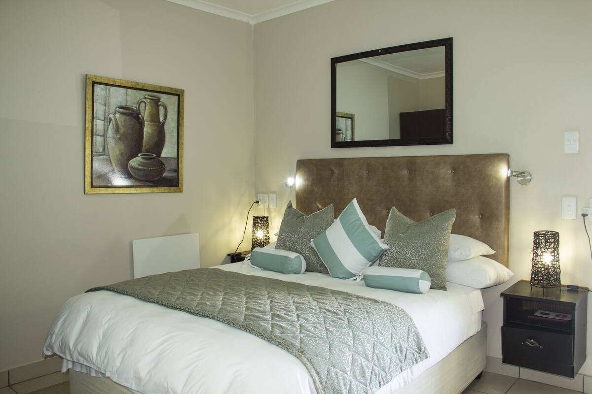 A neatly made bed with a patterned bedspread, a headboard, and multiple pillows. There is a bedside table with a lamp and a small drawer. A framed artwork hangs on the wall above the bed.